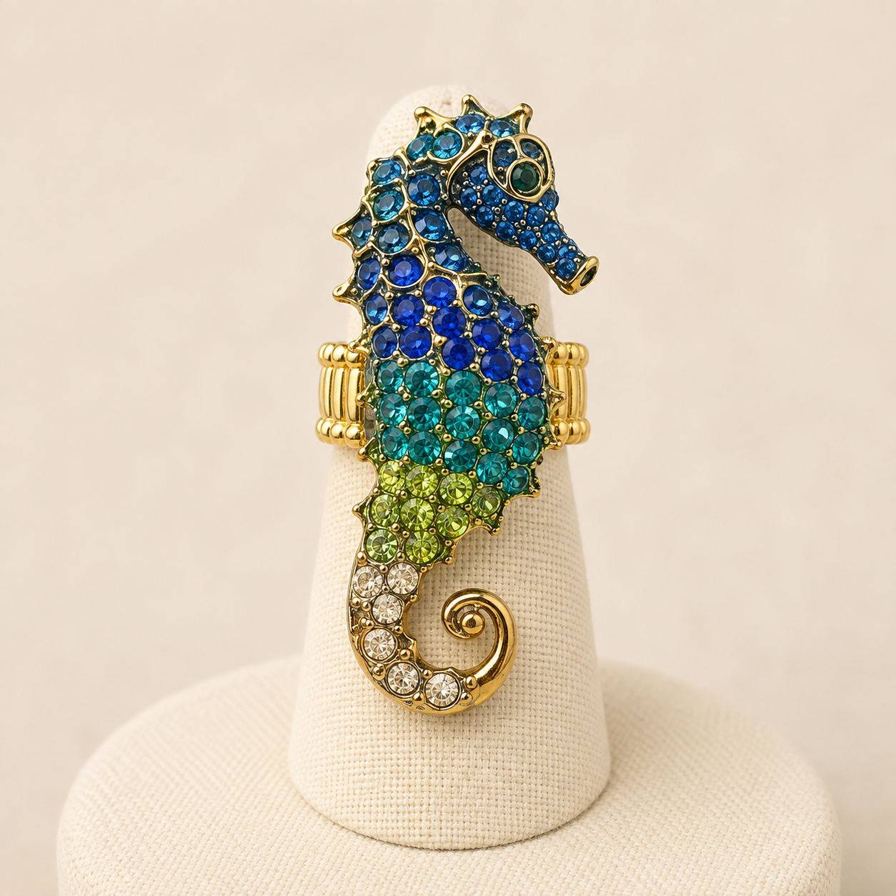 A gold seahorse ring embellished with blue, green, and turquoise rhinestones, displayed on a cream-colored jewelry cone stand against a warm beige background.