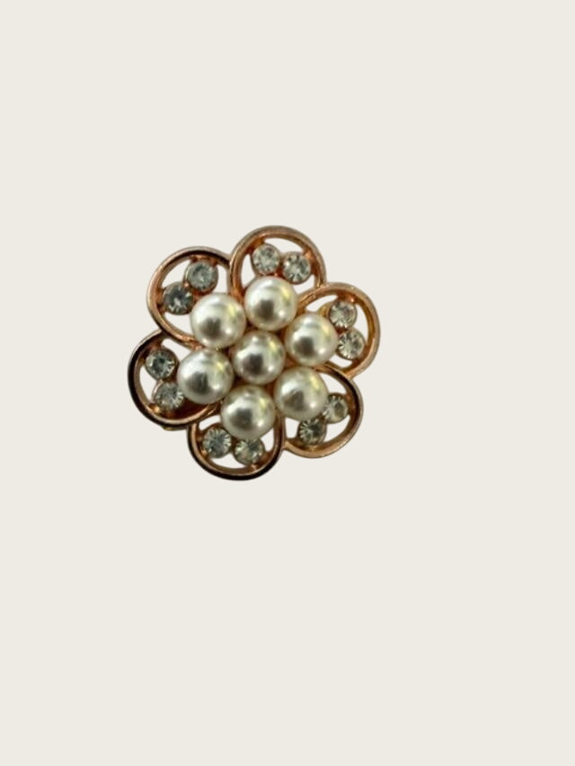 A vintage-style flower-shaped brooch featuring white faux pearls and clear rhinestones in a rose gold-tone setting, displayed on a textured gray background.