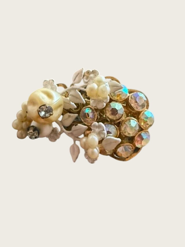 A vintage-style brooch featuring cream-colored faux pearls, white floral elements, and a cluster of faceted iridescent rhinestones in a gold-tone setting, resting on a dark mottled surface.