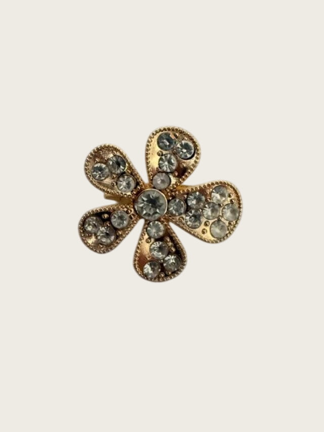 A gold-tone flower-shaped brooch embellished with clear rhinestones, resting on a textured white surface.