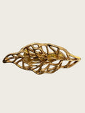 A gold-toned leaf-shaped brooch with an openwork, filigree design, displayed on a textured light gray surface.