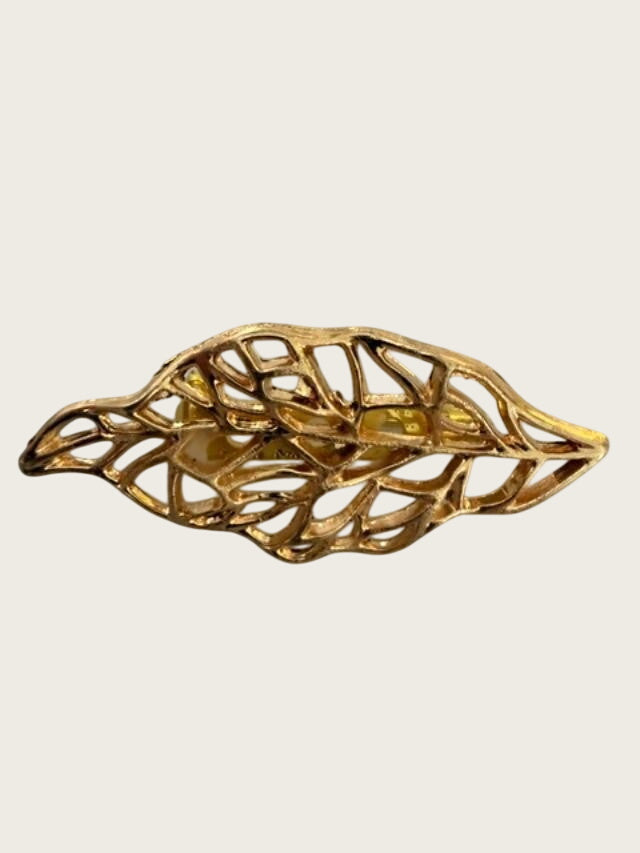 A gold-toned leaf-shaped brooch with an openwork, filigree design, displayed on a textured light gray surface.