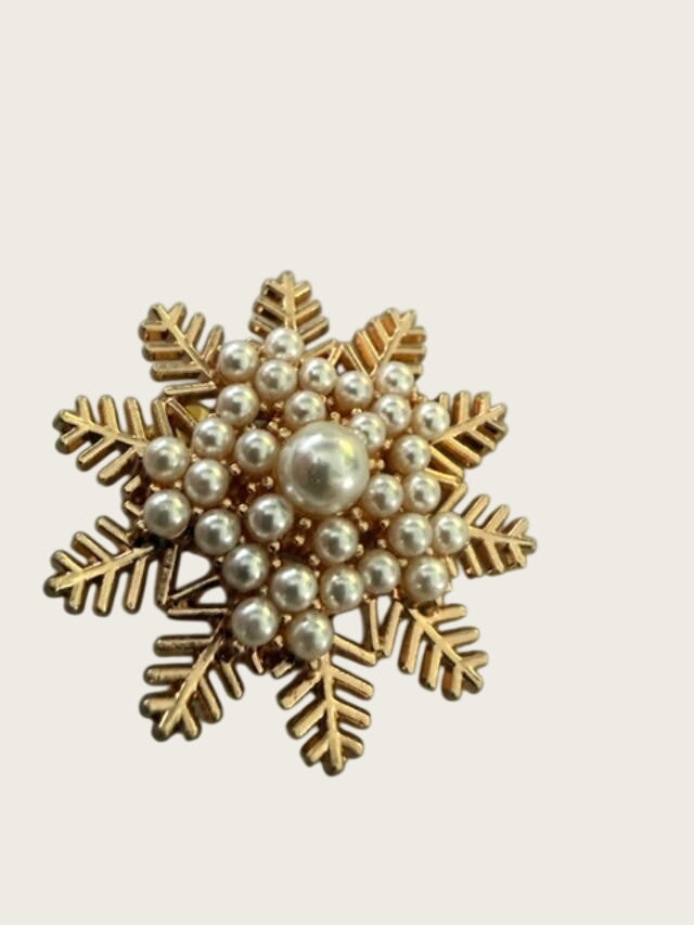 A snowflake-shaped brooch featuring a gold-tone metal frame with a central pearl and surrounding small white faux pearls, displayed on a textured gray background.